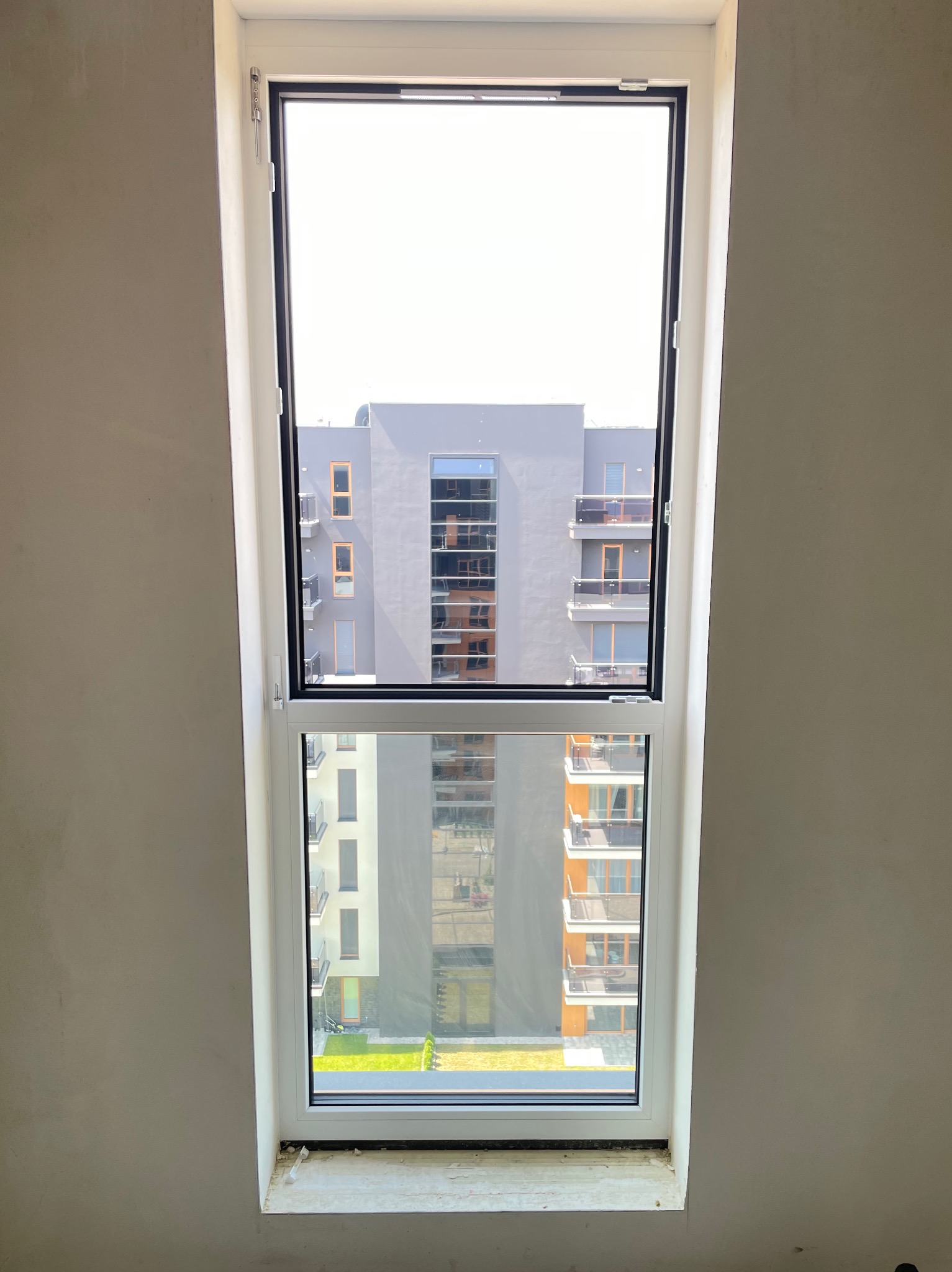Nowe białe okno z widokiem na budynek mieszkalny z balkonami, widoczne ślady po montażu na ścianie.