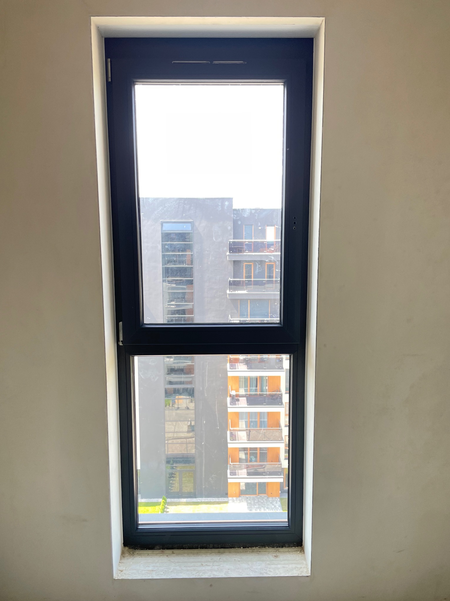 Wąskie, pionowe okno z czarną ramą, widok na nowoczesny budynek z balkonami, ściana w surowym stanie przed wykończeniem.