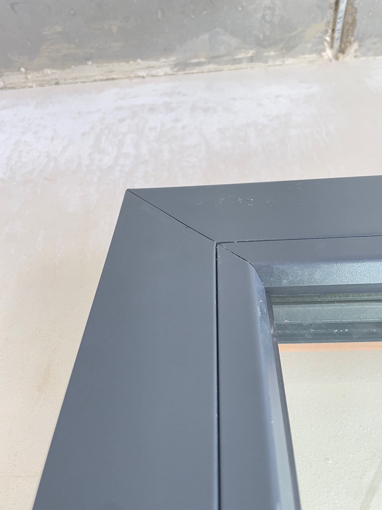 Szczegółowe zbliżenie na narożnik szarej ramy okiennej z widocznymi śladami niedawnego uszczelniania silikonem i surowym wykończeniem ściany w tle.