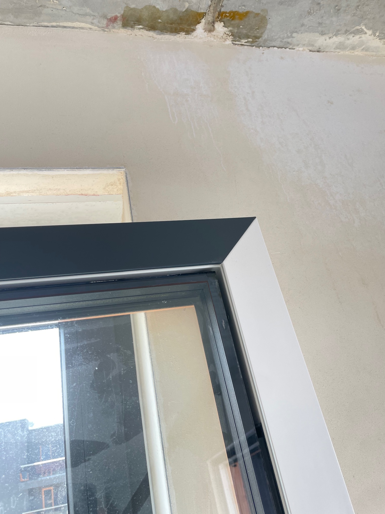 Fragment okna z czarną górną ramą i białą boczną ramą, widoczne zabrudzenia na szybie i surowy stan wykończenia ściany i sufitu.