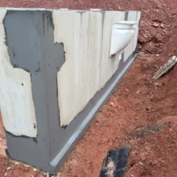 Betonowy fundament budynku z nałożoną izolacją przeciwwilgociową w kolorze szarym, widoczne rury odprowadzające wodę, wykop z czerwoną ziemią.