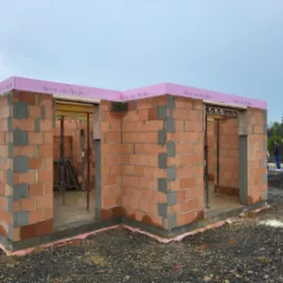 Wznoszone ściany domu z czerwonej cegły ceramicznej z widocznym szalunkiem i izolacją przeciwwilgociową, na zewnątrz rozsypany gruz.