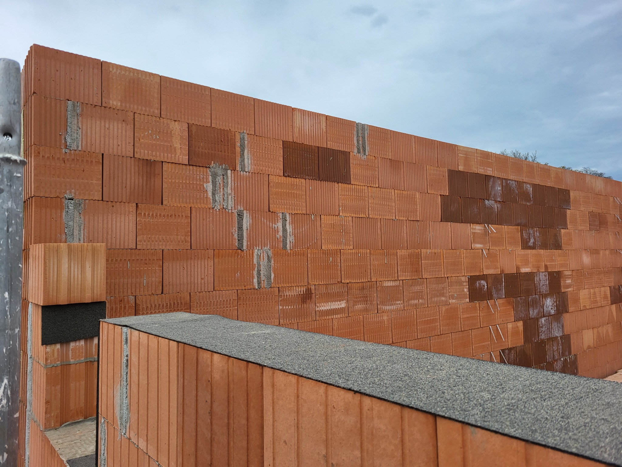 Wznoszona ściana zewnętrzna z czerwonych bloczków ceramicznych Porotherm, widoczne spoiny wypełnione zaprawą, fragment izolacji pionowej oraz szary parapet zewnętrzny.