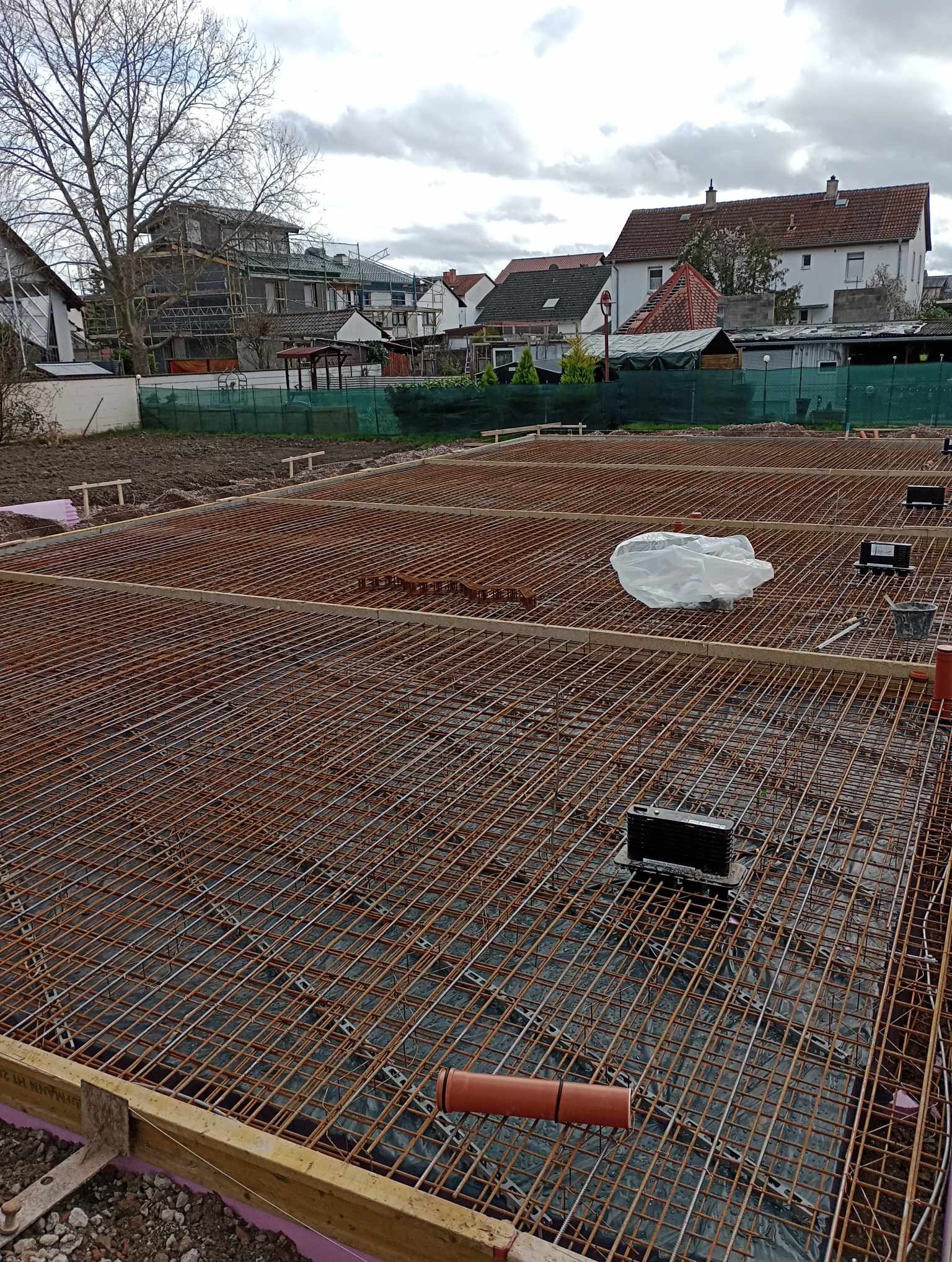 Zbrojenie fundamentu domu jednorodzinnego, widoczne pręty stalowe tworzące siatkę, elementy dystansowe z tworzywa, rury kanalizacyjne i folia izolacyjna, w tle zabudowa jednorodzinna i pochmurne...