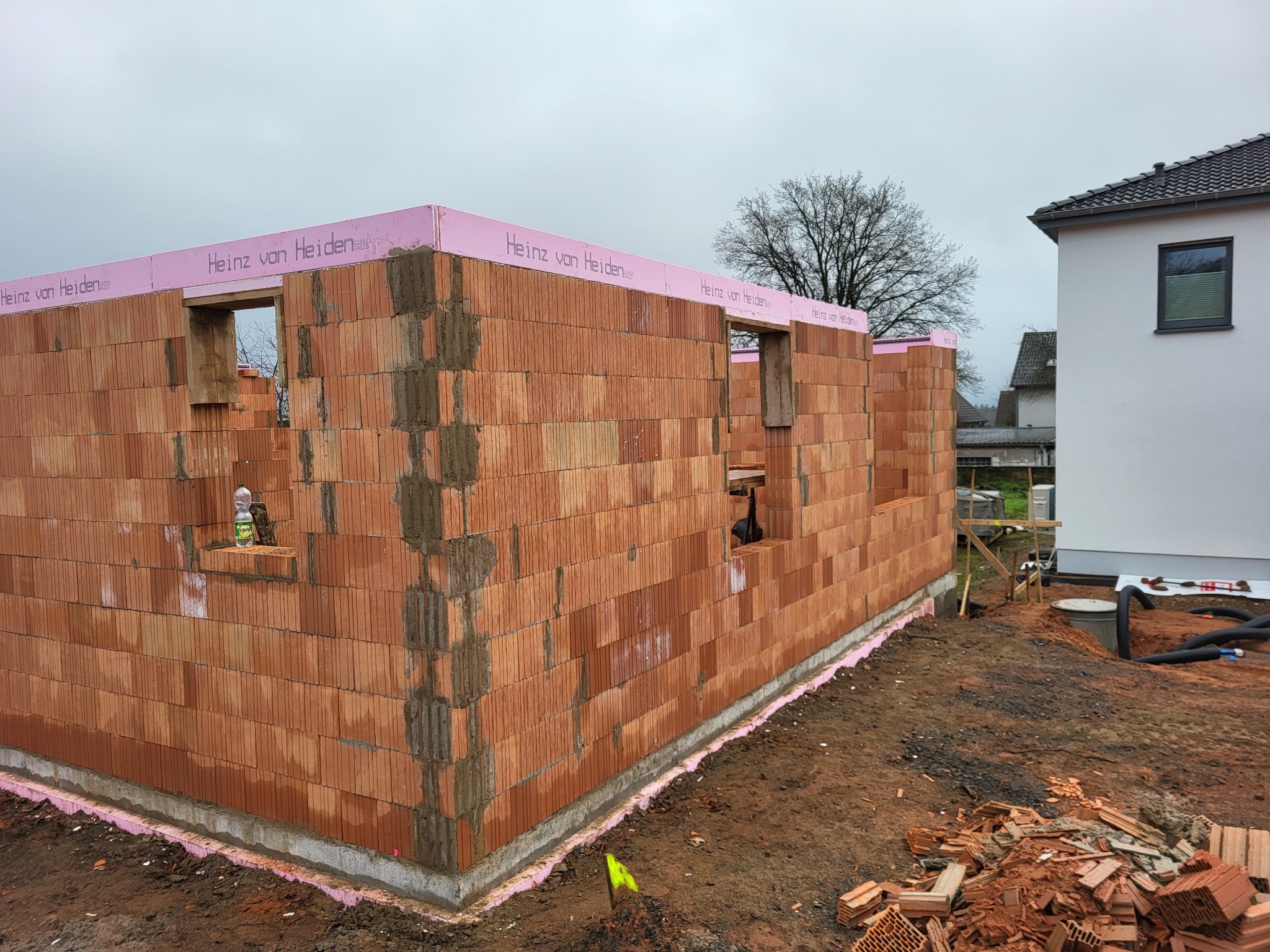 Wznoszenie ścian z czerwonej cegły ceramicznej z widocznymi otworami okiennymi, zabezpieczone różową izolacją, na fundamencie. Na parapecie butelka wody i figurka.