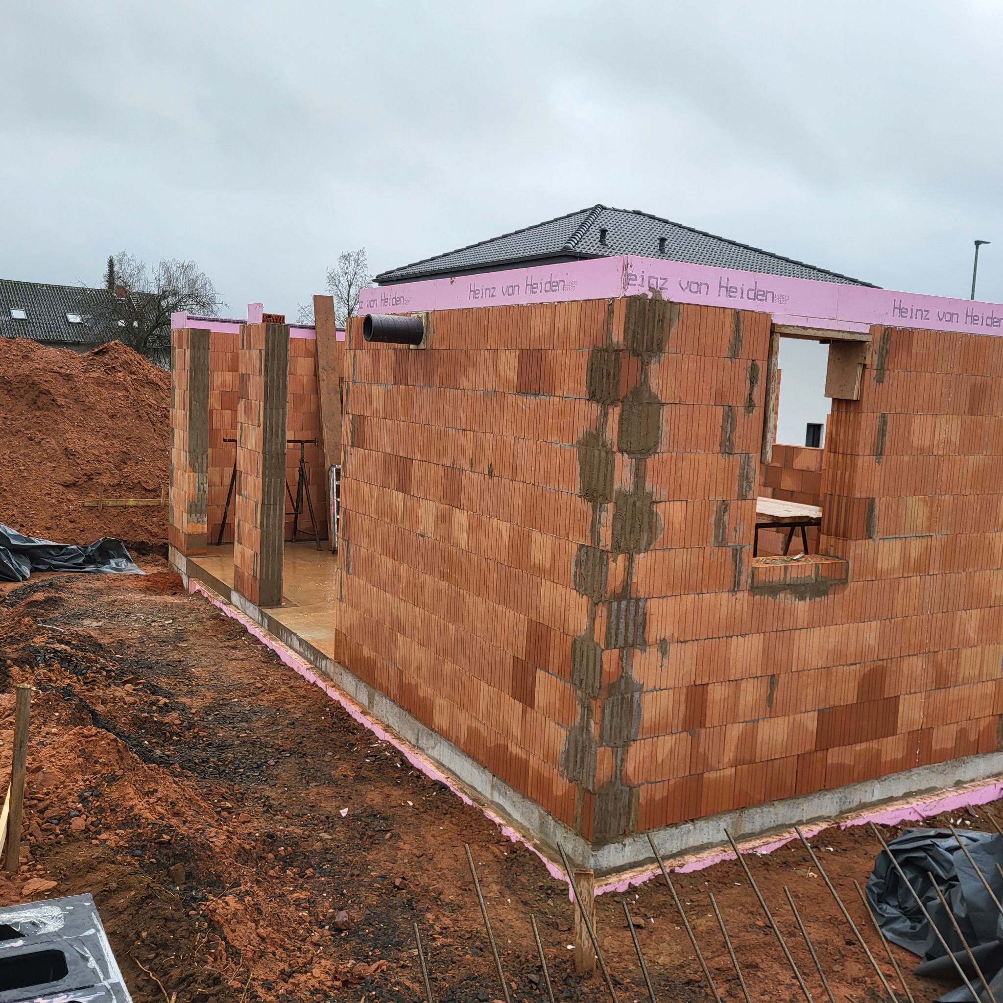 Wznoszenie ścian domu murowanego z czerwonej cegły ceramicznej. Widoczny fragment fundamentu zabezpieczony różową izolacją termiczną Heinz von Heiden. Na zewnątrz sterta ziemi i pręty zbrojeniowe...