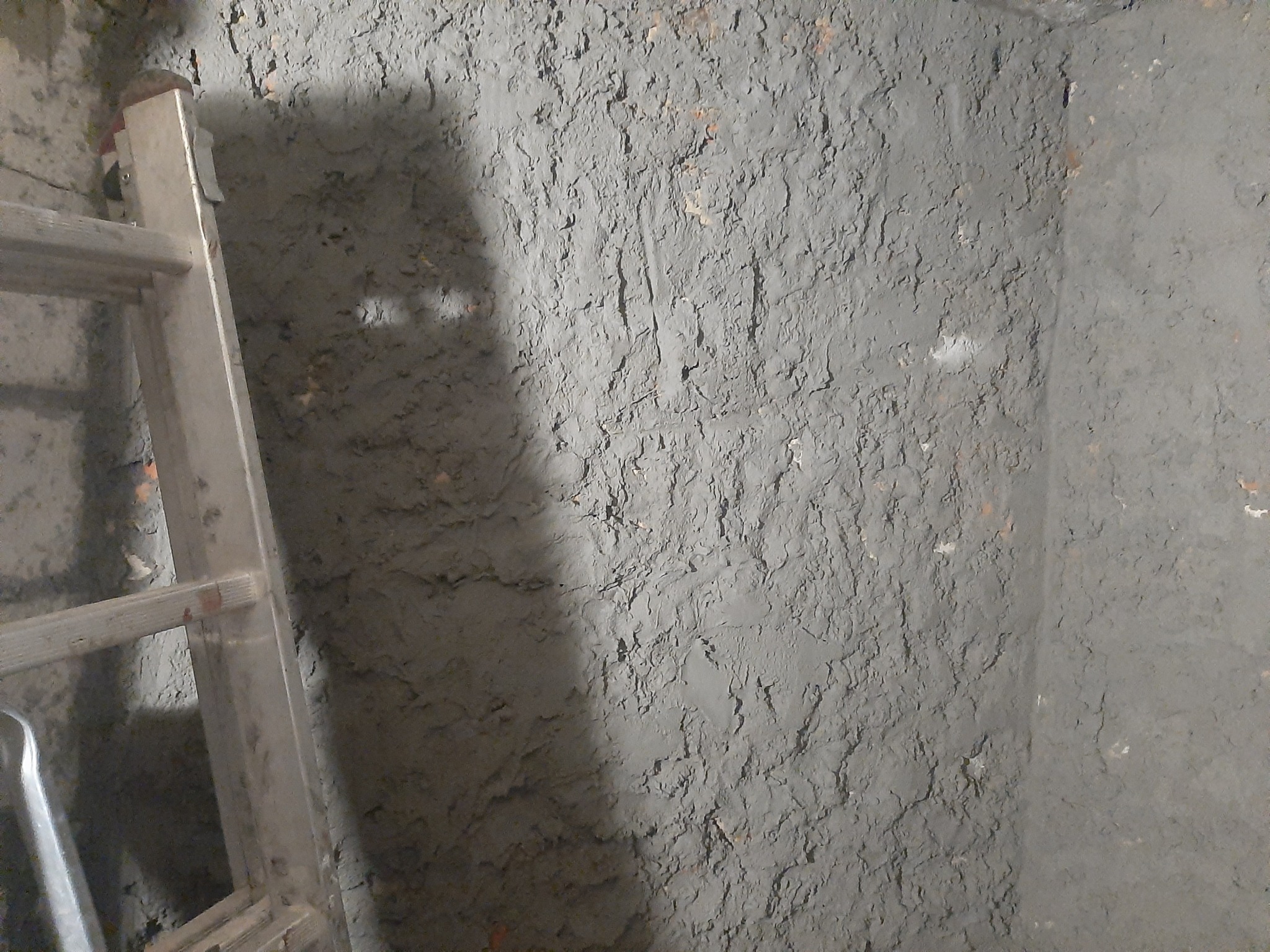 Szara, nierówna ściana z widoczną fakturą i fragmentem drabiny aluminiowej po lewej stronie.