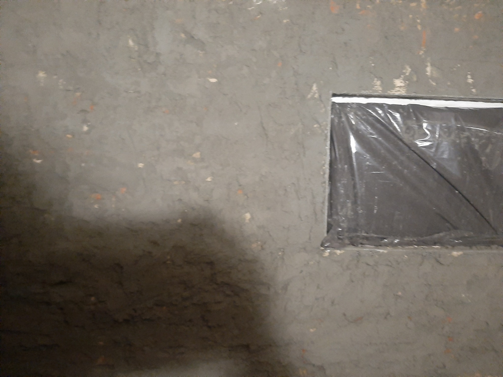 Surowa, szara ściana z niedokończonym tynkiem i zakrytym folią otworem okiennym.