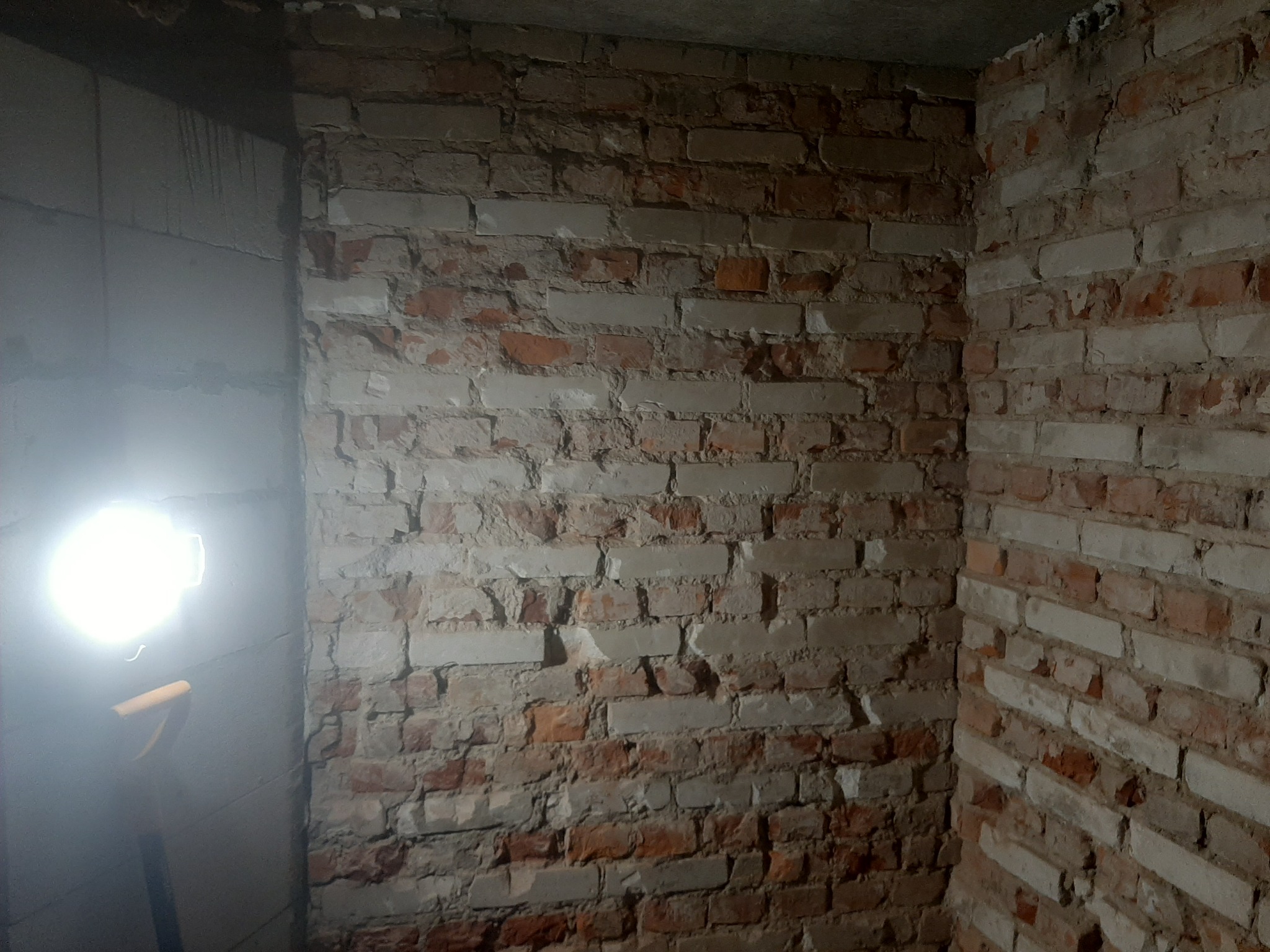 Fragment wnętrza z surowymi, częściowo otynkowanymi ścianami z czerwonej cegły, oświetlony mocnym światłem lampy roboczej, z widocznym fragmentem szpadla w lewym dolnym rogu.