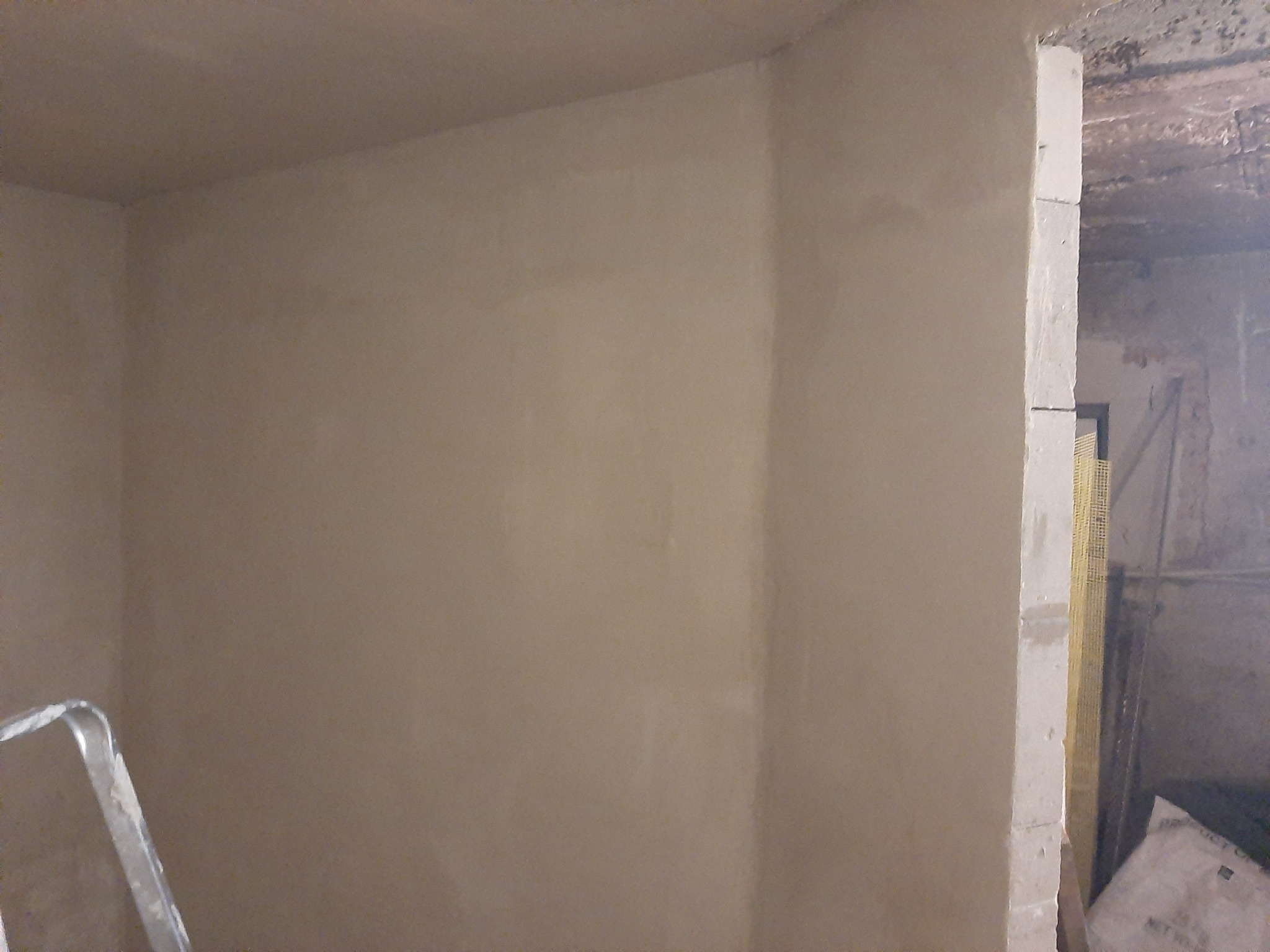 Wnętrze w trakcie prac wykończeniowych: świeżo otynkowana ściana w odcieniach beżu, widoczne ślady po łatach, fragment drabiny malarskiej w lewym dolnym rogu oraz mur z pustaków z siatką...