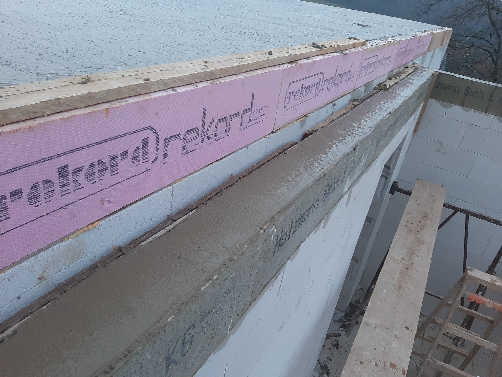 Wznoszenie ściany z betonu komórkowego z widocznym wieńcem zbrojonym, ociepleniem styropianem różowym i szalunkiem drewnianym.