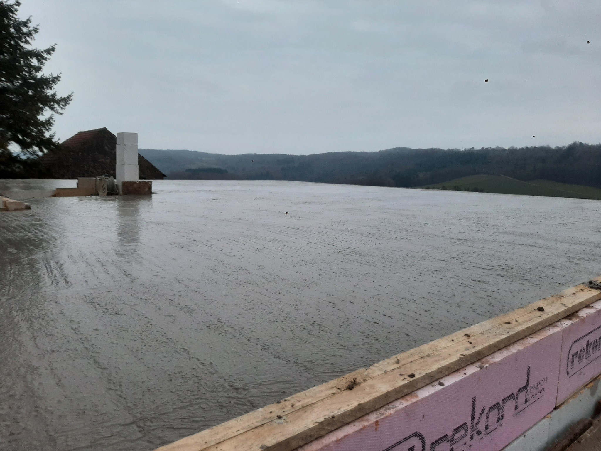 Wylany fundament z widocznym szalunkiem i izolacją termiczną, w tle zalesione wzgórza i pochmurne niebo.