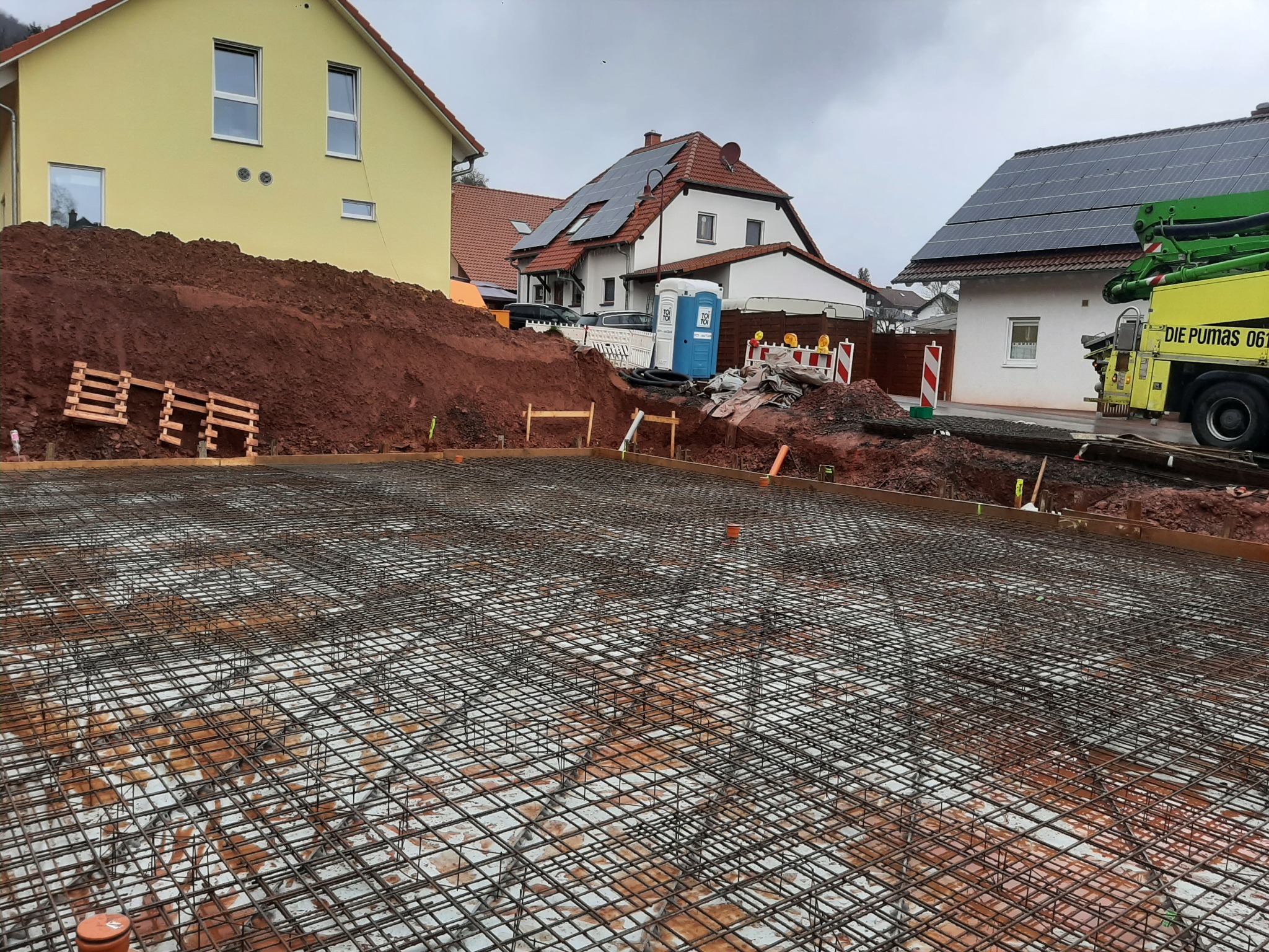 Zbrojenie fundamentu przygotowane do zalania betonem, widoczna siatka stalowa, szalunki i fragment pompy do betonu na tle zabudowy jednorodzinnej.