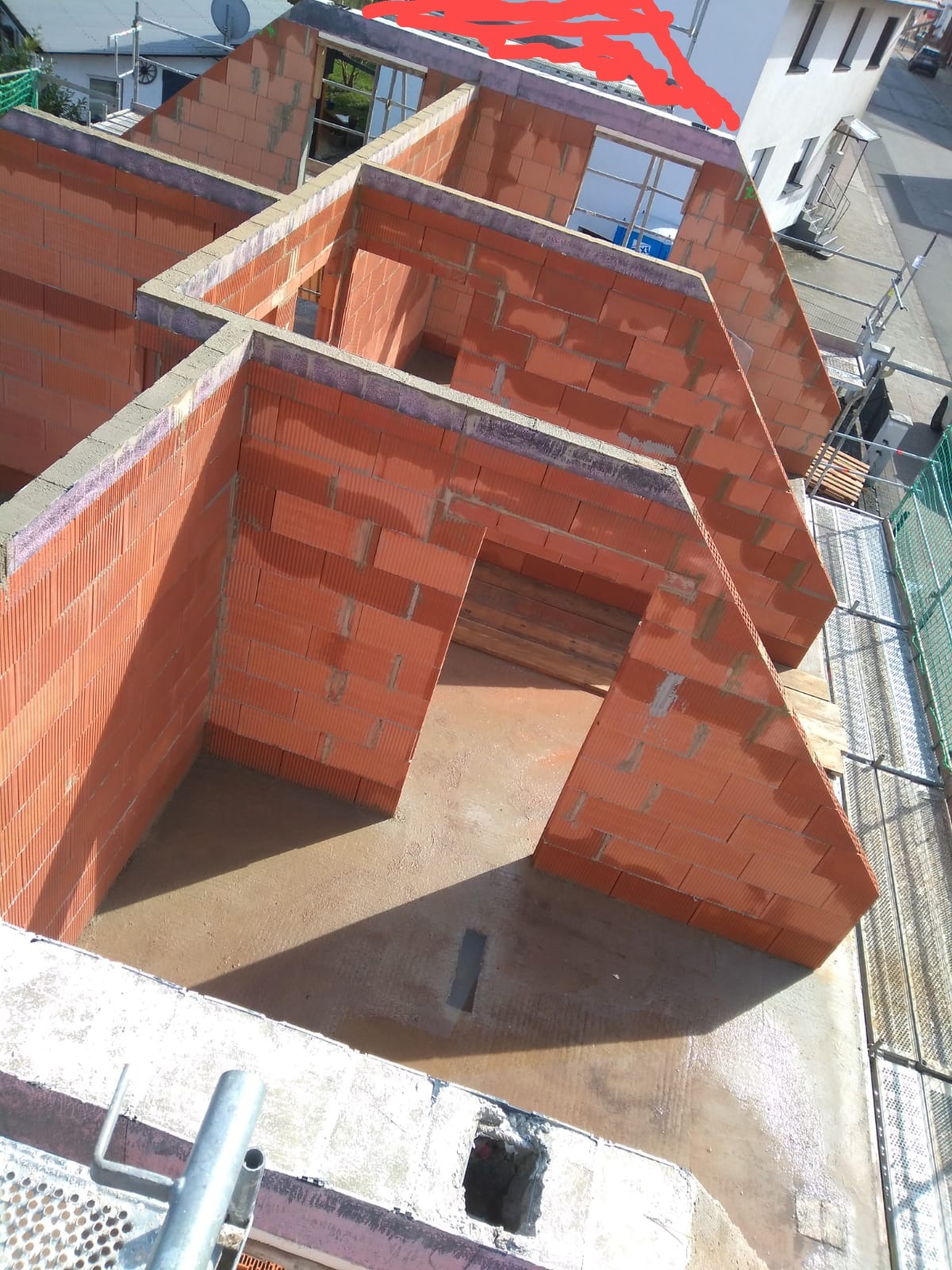 Wznoszenie ścian zewnętrznych domu z czerwonej cegły ceramicznej, widok z góry na częściowo ukończoną konstrukcję z otworami okiennymi i drzwiowymi, z widocznymi elementami rusztowania.