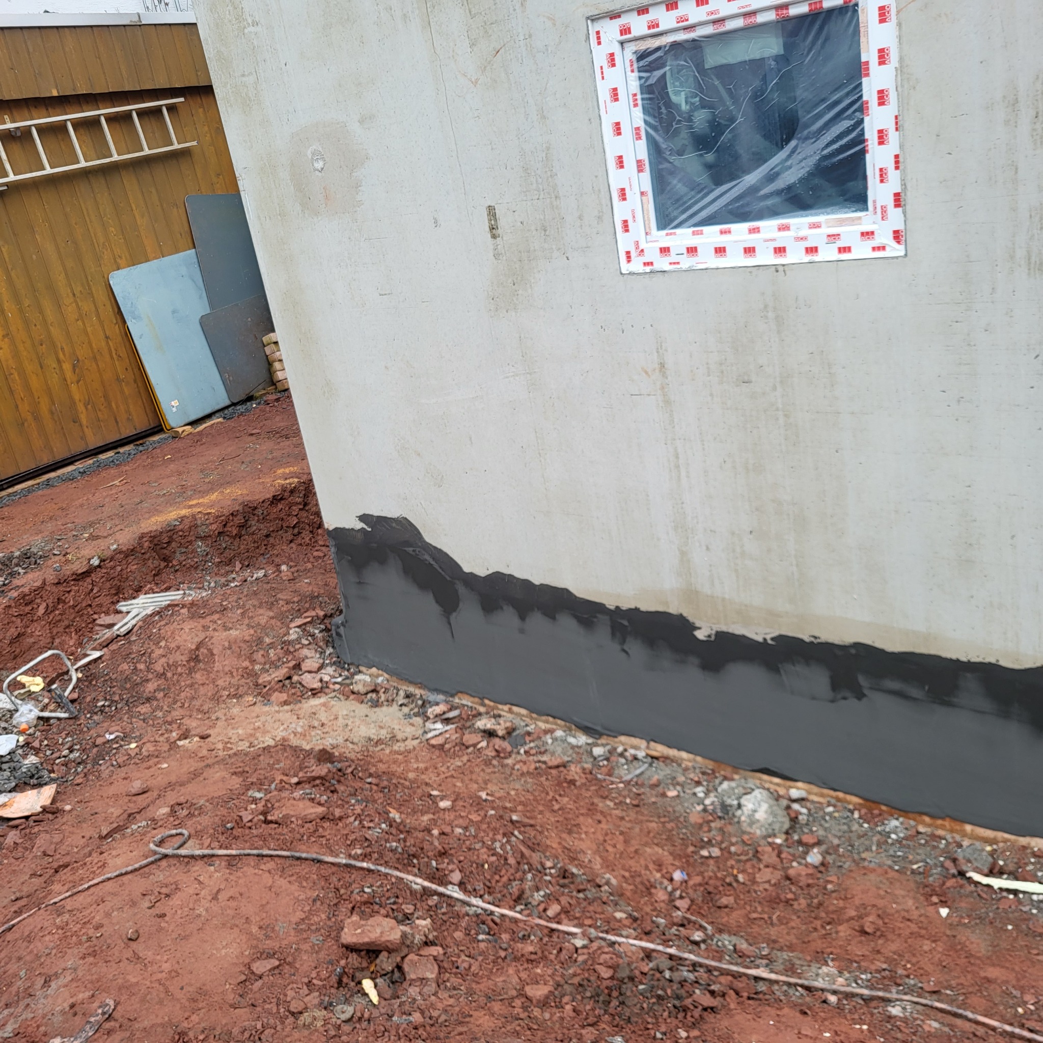 Szara ściana budynku w trakcie budowy z oknem zabezpieczonym folią, widoczne zaizolowane fundamenty i ziemia wokół