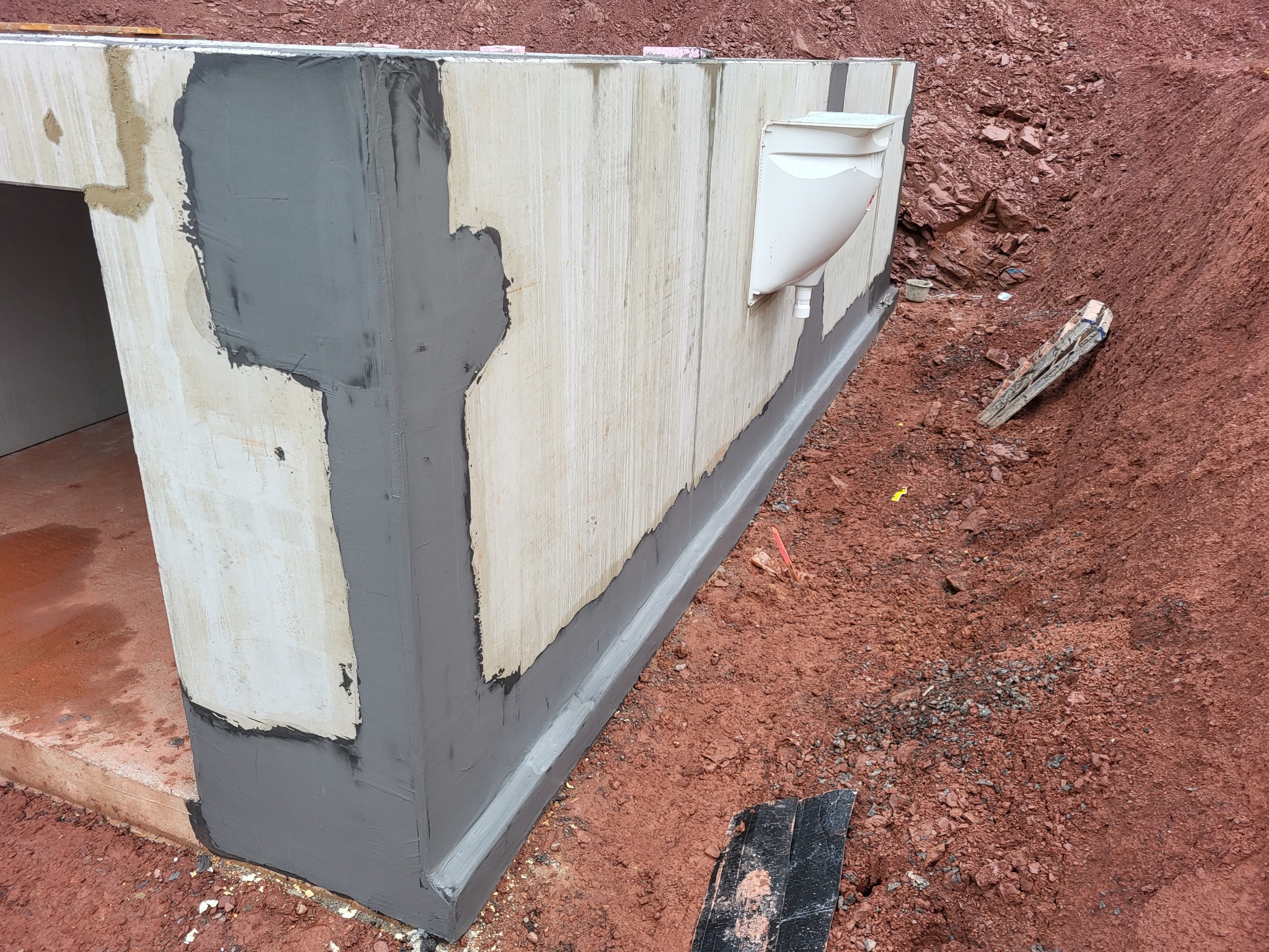 Betonowy fundament budynku z nałożoną izolacją przeciwwilgociową w kolorze szarym, widoczne rury odprowadzające wodę, wykop z czerwoną ziemią.