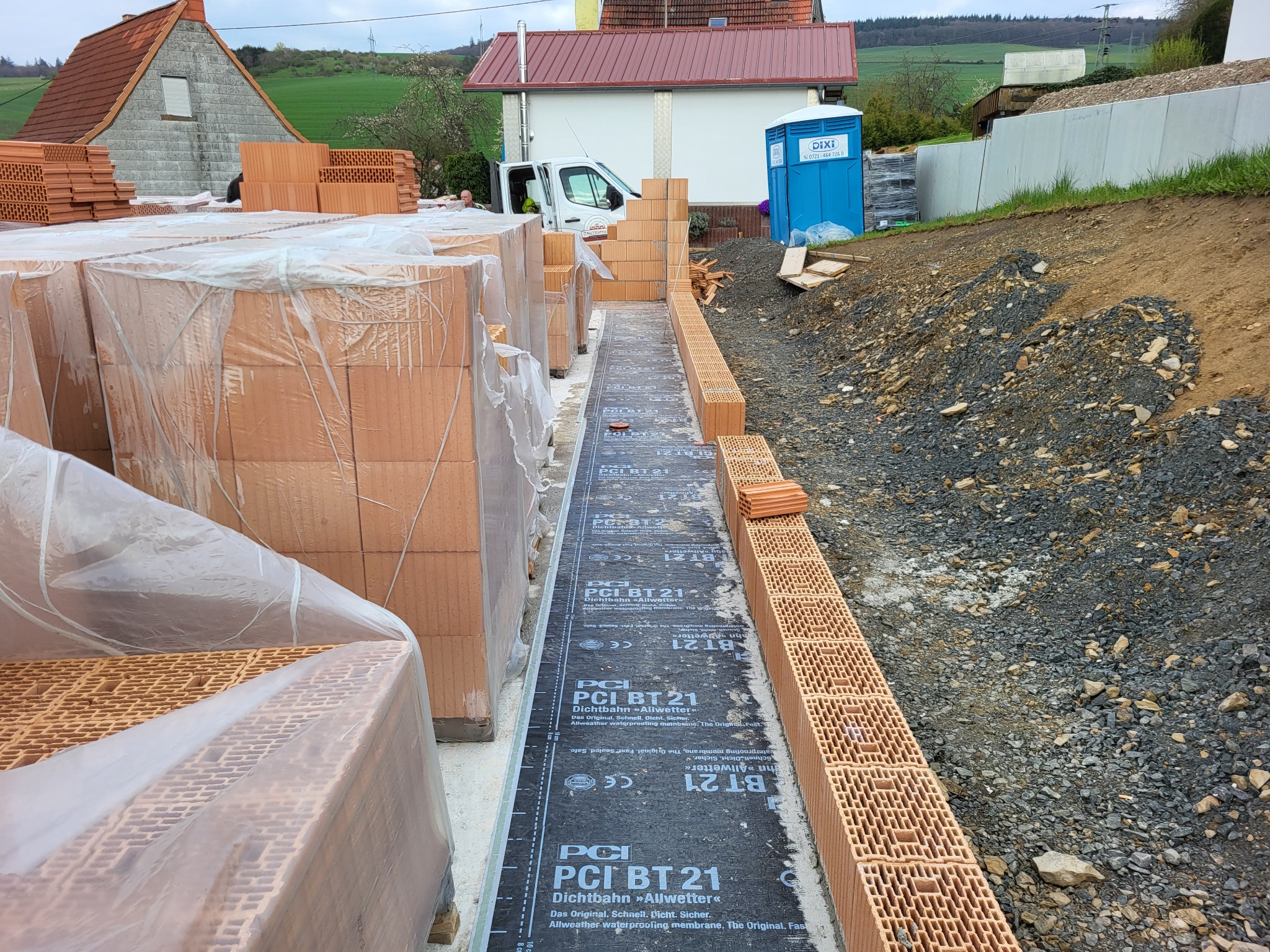 Wznoszenie muru z pustaków ceramicznych na fundamencie zabezpieczonym czarną membraną izolacyjną PCI BT 21, widoczne palety z pustakami w folii, w tle budynek gospodarczy i przenośna toaleta.