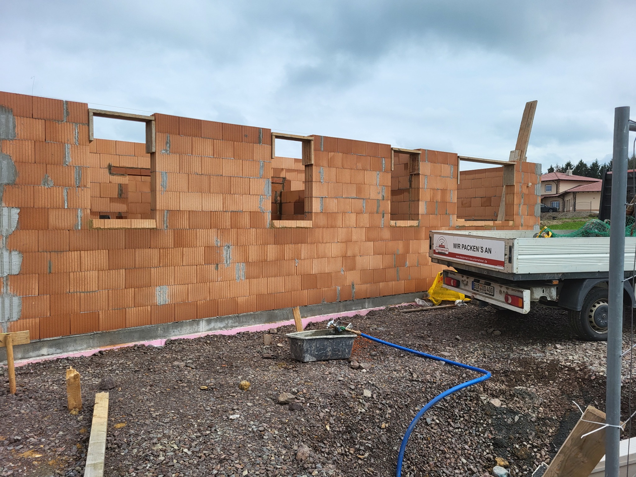 Wznoszenie muru z czerwonej cegły ceramicznej porotherm z otworami, widoczne otwory okienne z drewnianymi nadprożami, na pierwszym planie gruz i wąż doprowadzający wodę, w tle samochód dostawczy...