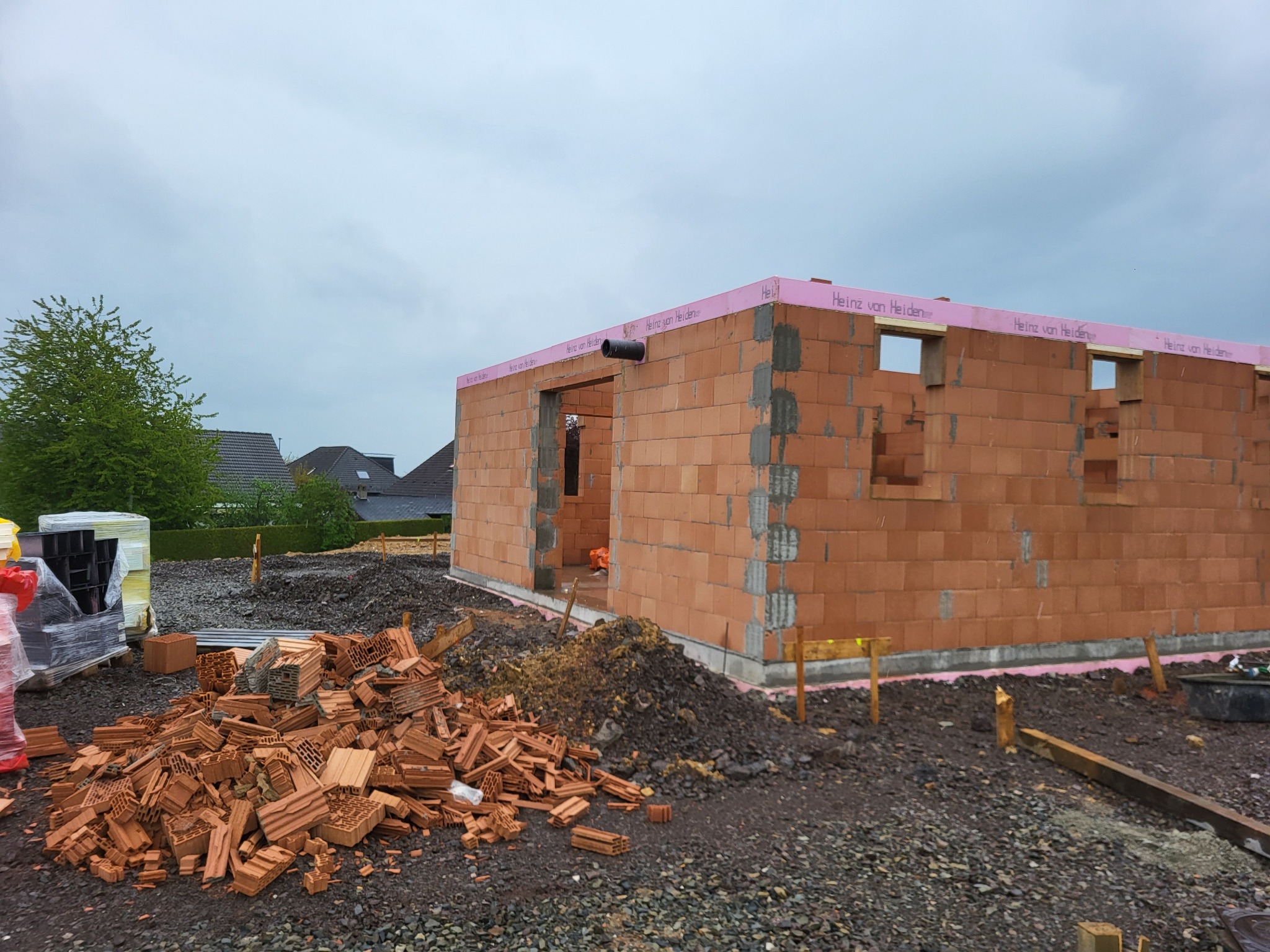 Wznoszenie ścian parterowego budynku z czerwonej cegły ceramicznej, widoczne otwory okienne i drzwiowe, zabezpieczone różową pianką izolacyjną, na pierwszym planie sterta potłuczonych cegieł.