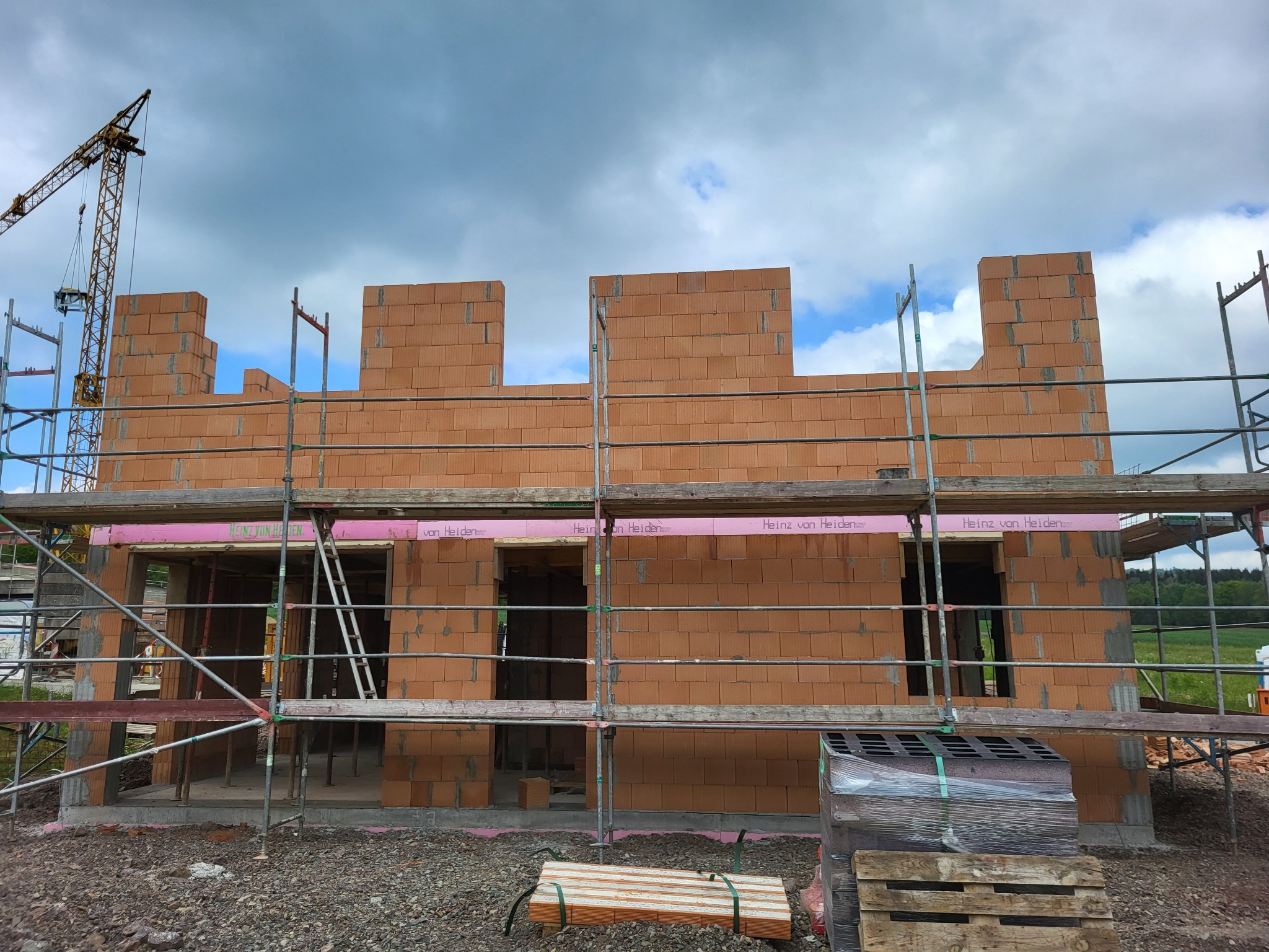 Wznoszenie ścian zewnętrznych domu z czerwonej cegły ceramicznej, widoczne rusztowania, dźwig w tle i przygotowane materiały budowlane.