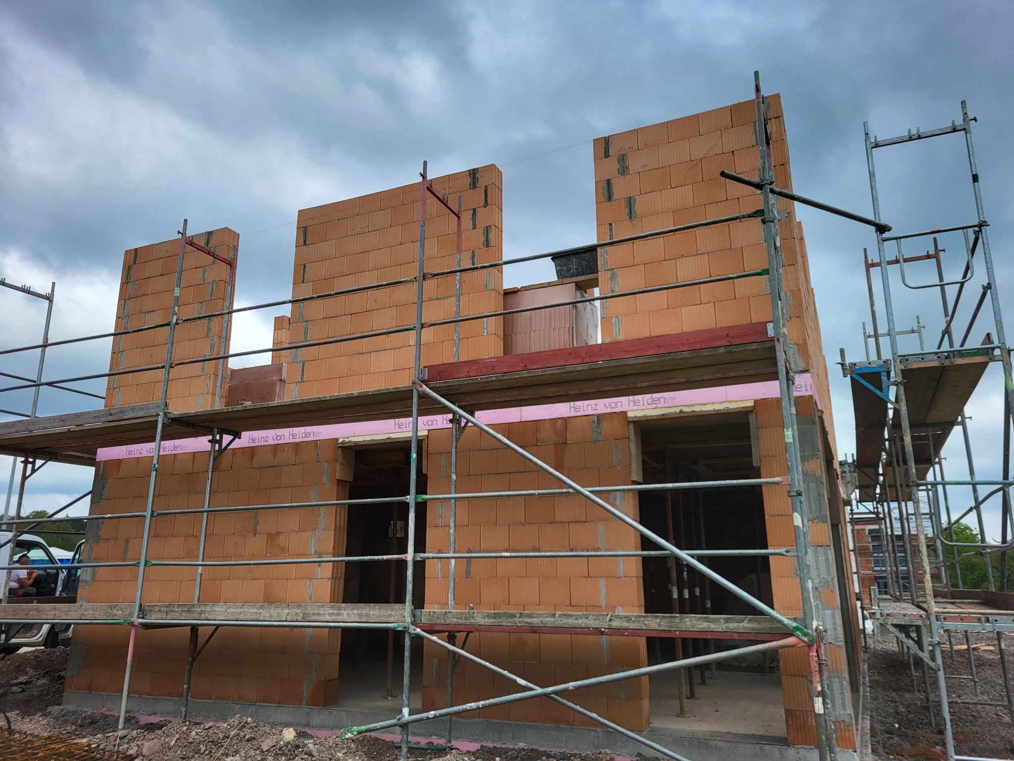 Surowa konstrukcja domu z czerwonej cegły ceramicznej, widoczne rusztowania i elementy izolacji termicznej z logo producenta Heinz von Heiden, pochmurne niebo w tle.