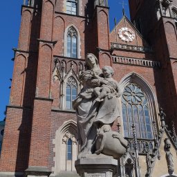 fotopol - Statua Matki Boskiej z Dzieciątkiem na tle ceglanej fasady gotyckiej katedry z zegarem słonecznym i ozdobnymi oknami.