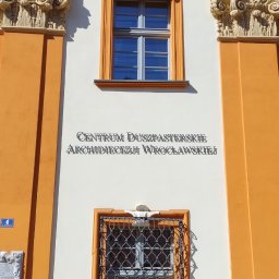 fotopol - Fasada budynku Centrum Duszpasterskiego Archidiecezji Wrocławskiej z dekoracyjnym herbem, oknami z kratami i pomarańczowymi akcentami architektonicznymi.