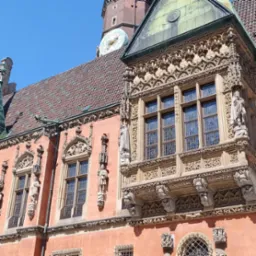 Fasada zabytkowej kamienicy z bogato zdobionym balkonem, rzeźbami i zegarem wieżowym w tle.