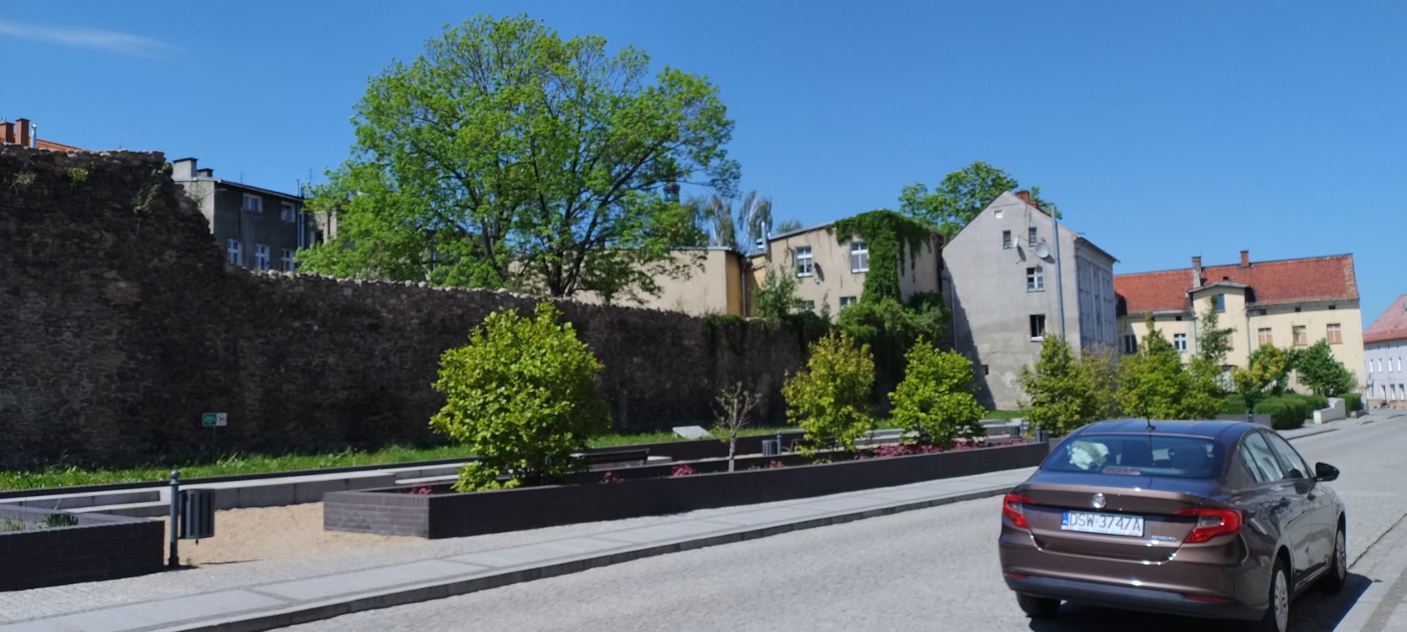 Ulica z odnowionym murem, ozdobnymi rabatami z zielenią i brązowym samochodem osobowym w słoneczny dzień.