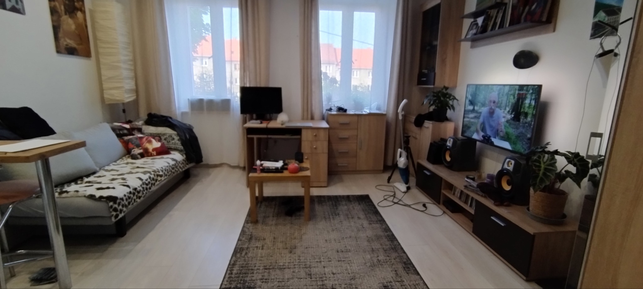 Widok na wnętrze pokoju dziennego z sofą z narzutą w panterkę, biurkiem z monitorem, komodą, dywanem w odcieniach szarości i szafką RTV z telewizorem i głośnikami.