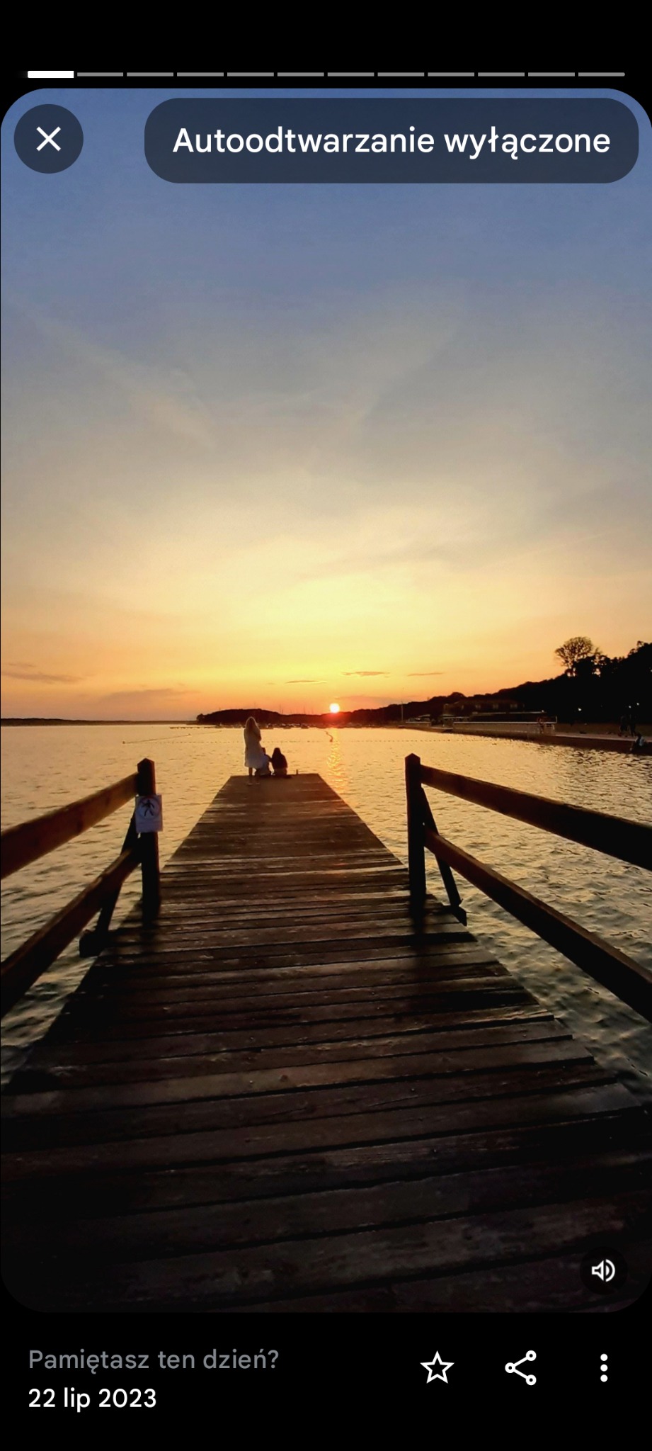 Pomost z drewnianą barierką prowadzący do jeziora o zachodzie słońca, sylwetki dwóch osób na końcu pomostu.