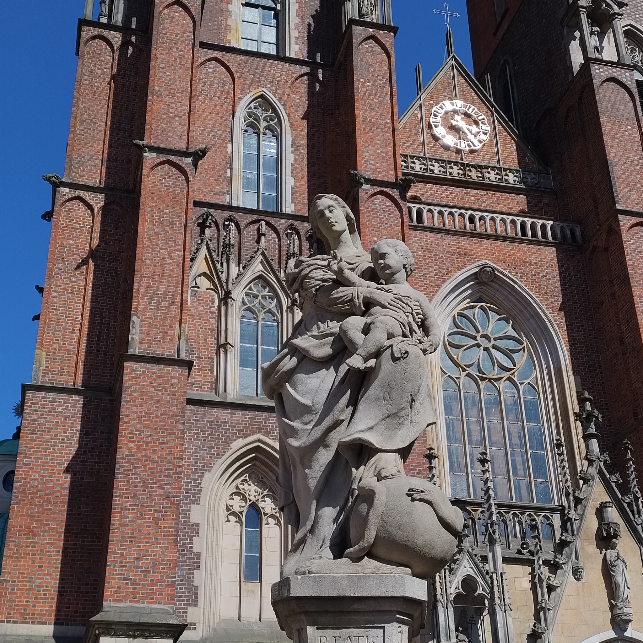 Statua Matki Boskiej z Dzieciątkiem na tle ceglanej fasady gotyckiej katedry z zegarem słonecznym i ozdobnymi oknami.