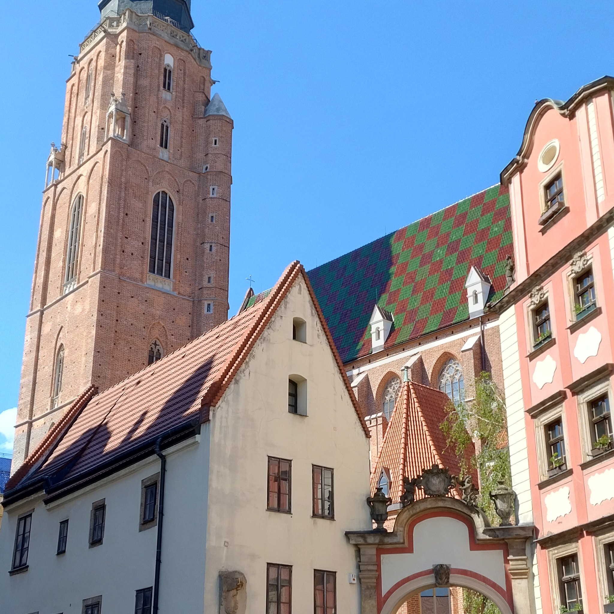 Fragment architektury miejskiej: wieża kościoła z czerwonej cegły, kamienice z dachami o różnej kolorystyce (czerwona dachówka, zielono-czerwona szachownica), detale architektoniczne.