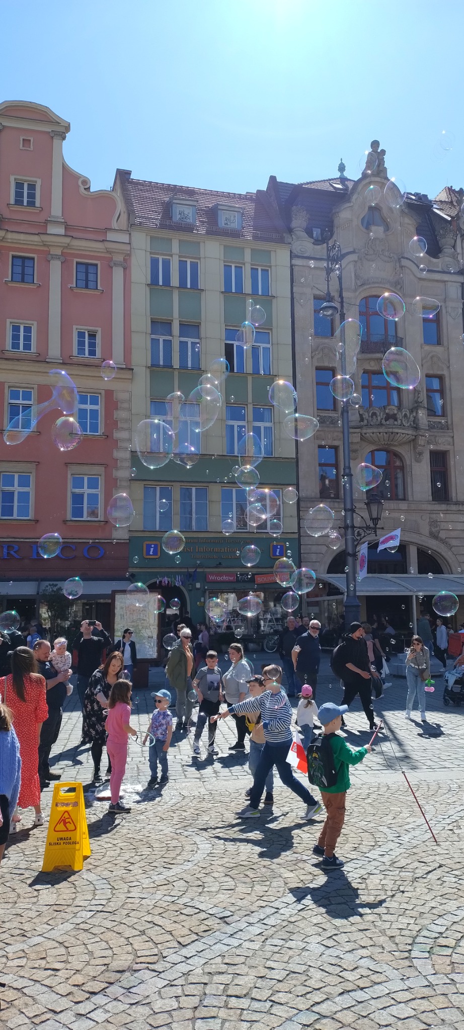 Rynek miejski z kolorowymi kamienicami, tłum ludzi obserwujących puszczanie baniek mydlanych, żółty znak ostrzegawczy o śliskiej podłodze w dolnym lewym rogu.