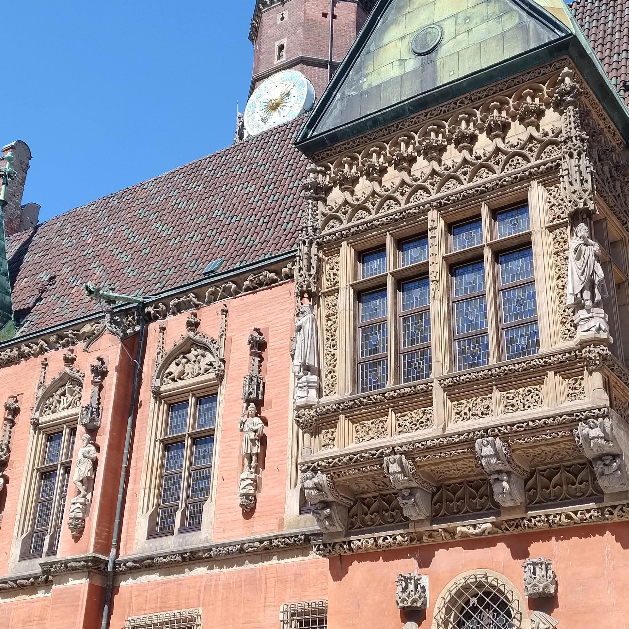 Fasada zabytkowej kamienicy z bogato zdobionym balkonem, rzeźbami i zegarem wieżowym w tle.