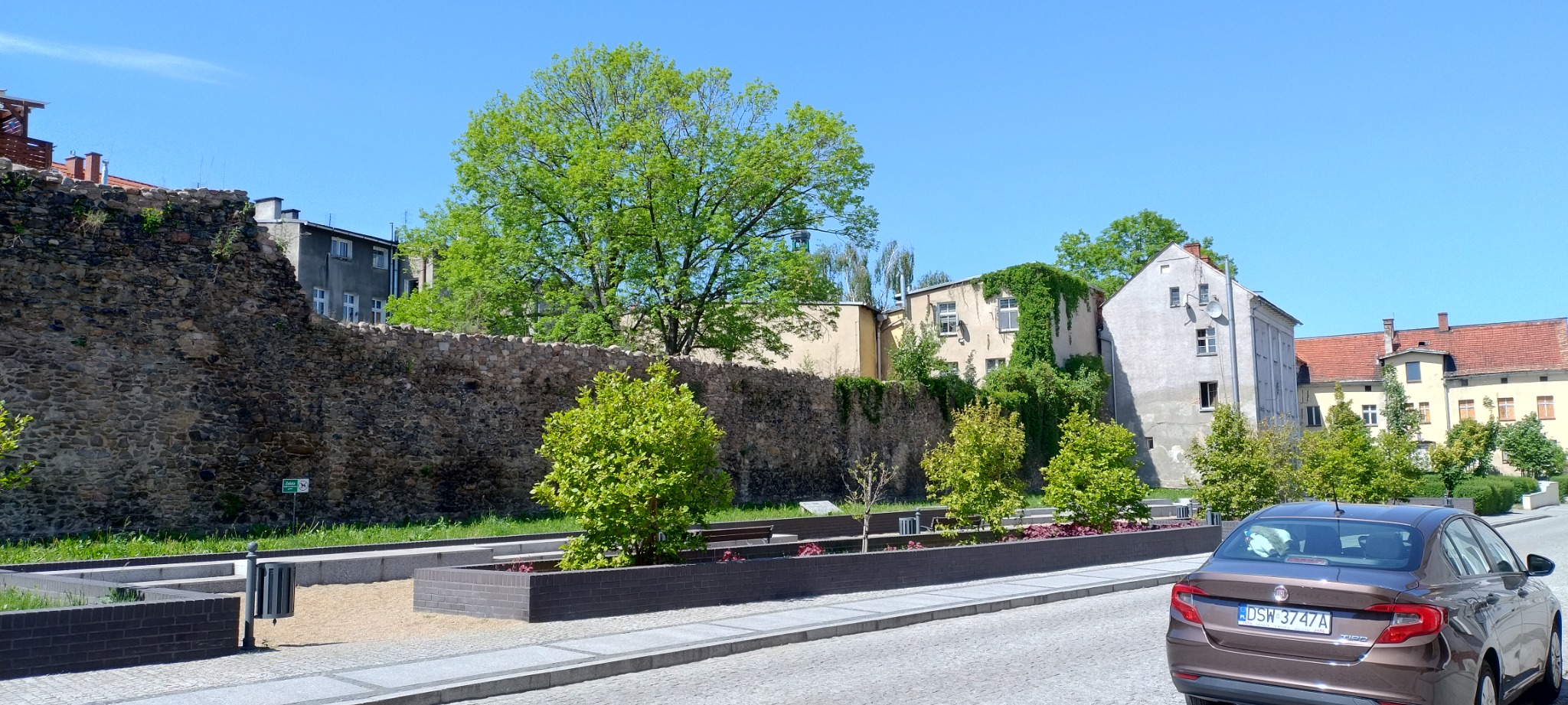 Fragment miejskiego krajobrazu z brukowaną drogą, fragmentem kamiennego muru, niską zielenią i budynkami w tle, z brązowym samochodem w prawym dolnym rogu.