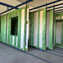 JK HAUS-WIĄZAR-SYSTEM SPÓŁKA Z OGRANICZONĄ ODPOWIEDZIALNOŚCIĄ - Elementy ścian z zielonej kantówki z oknami i drzwiami, przygotowane do montażu, ustawione na szynach w hali produkcyjnej.