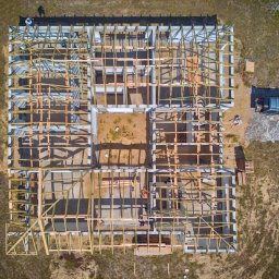 JK HAUS-WIĄZAR-SYSTEM SPÓŁKA Z OGRANICZONĄ ODPOWIEDZIALNOŚCIĄ - Widok z drona na fundamenty i drewnianą konstrukcję ścian powstającego domu, z zaparkowanym samochodem i widoczną osobą na placu budowy.