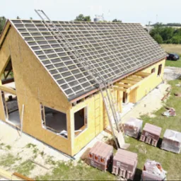 Wznoszenie konstrukcji domu z drewnianych paneli, widoczna więźba dachowa z membraną, palety z dachówkami na trawniku, drabina oparta o dach, samochód w tle.