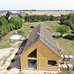 JK HAUS-WIĄZAR-SYSTEM SPÓŁKA Z OGRANICZONĄ ODPOWIEDZIALNOŚCIĄ - Wznoszenie konstrukcji budynku z drewnianym szkieletem i widoczną więźbą dachową, ujęcie z góry na tle wiejskiego krajobrazu z polami i zabudowaniami.