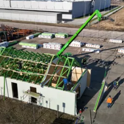 Montaż konstrukcji domu z prefabrykatów na betonowej płycie, widoczny dźwig i pracownicy podczas pracy na wysokościach.