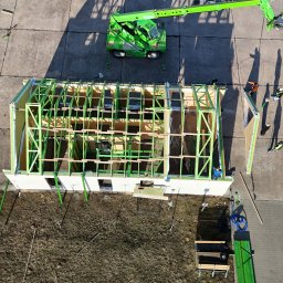 JK HAUS-WIĄZAR-SYSTEM SPÓŁKA Z OGRANICZONĄ ODPOWIEDZIALNOŚCIĄ - Budowa domu szkieletowego z zieloną konstrukcją, widok z góry z dźwigiem i pracownikami w kaskach, materiały budowlane obok