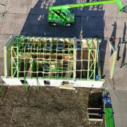 Budowa domu szkieletowego z zieloną konstrukcją, widok z góry z dźwigiem i pracownikami w kaskach, materiały budowlane obok