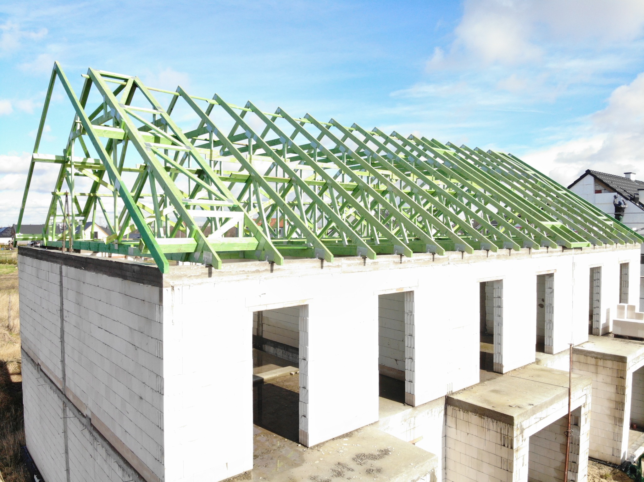 Wznoszenie konstrukcji dachu z zielonej więźby na budynku z białych bloczków, widok z góry na tle błękitnego nieba.