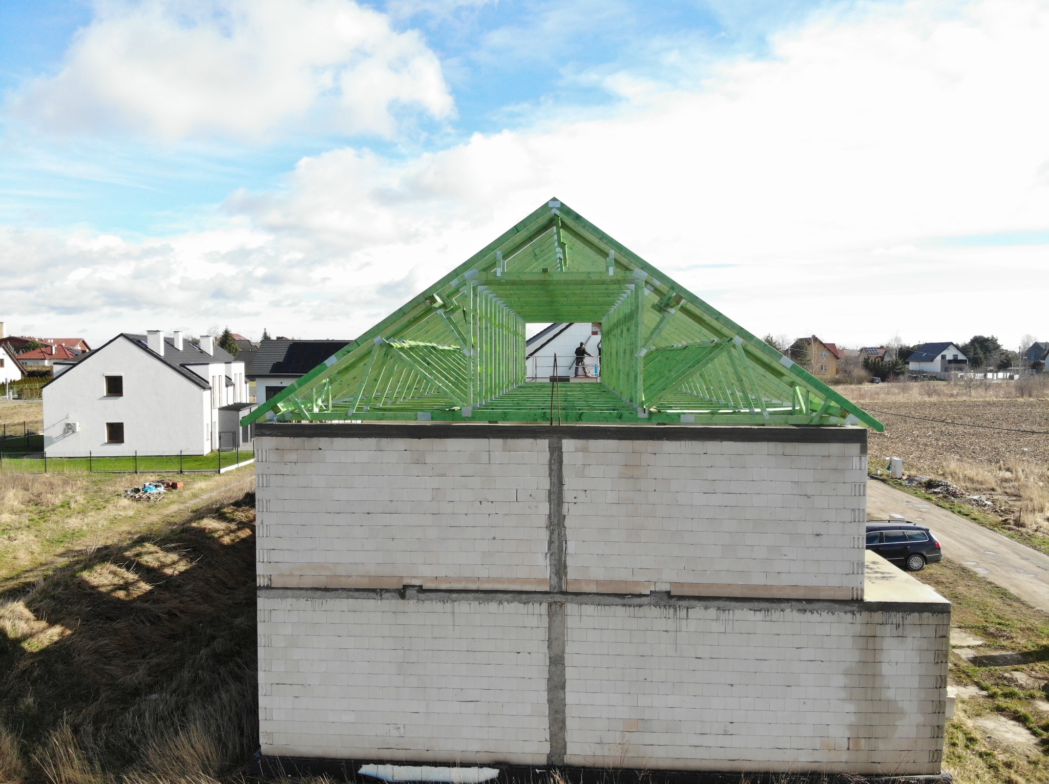 Wznoszenie konstrukcji dachu z impregnowanego drewna na budynku z białych bloczków, widoczny pracownik na górze, w tle zabudowa jednorodzinna i pole.