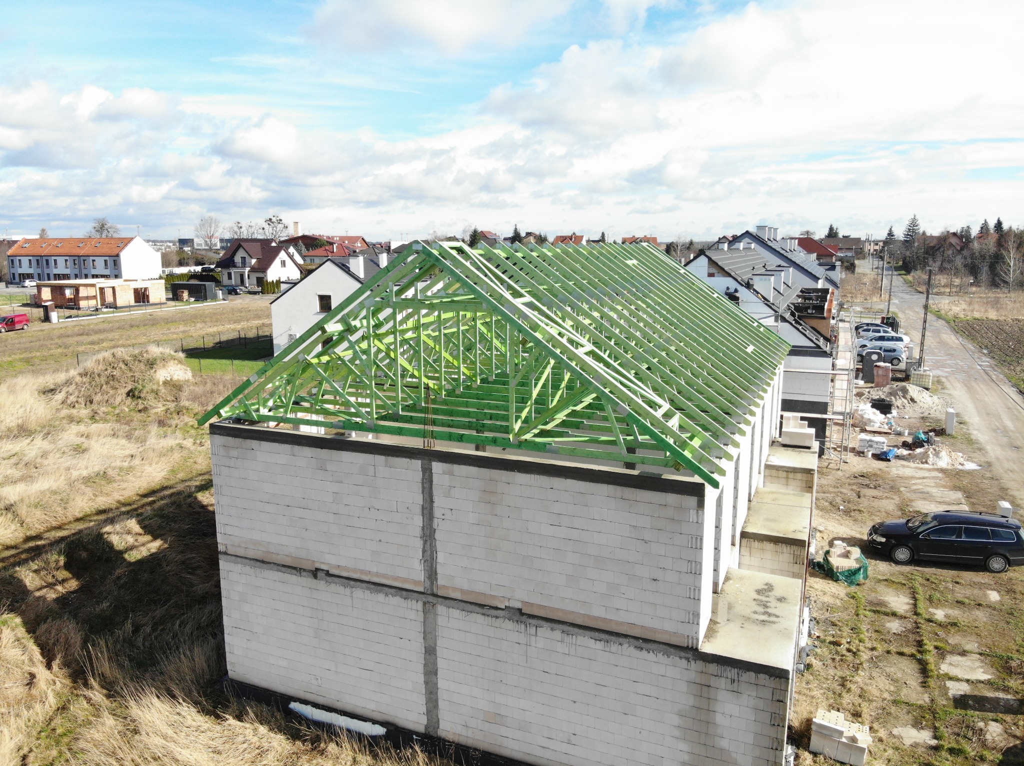 Wznoszenie dwukondygnacyjnego budynku z betonu komórkowego z widoczną, świeżo zaimpregnowaną, zieloną konstrukcją dachu, na tle zabudowy jednorodzinnej i błękitnego nieba z chmurami.