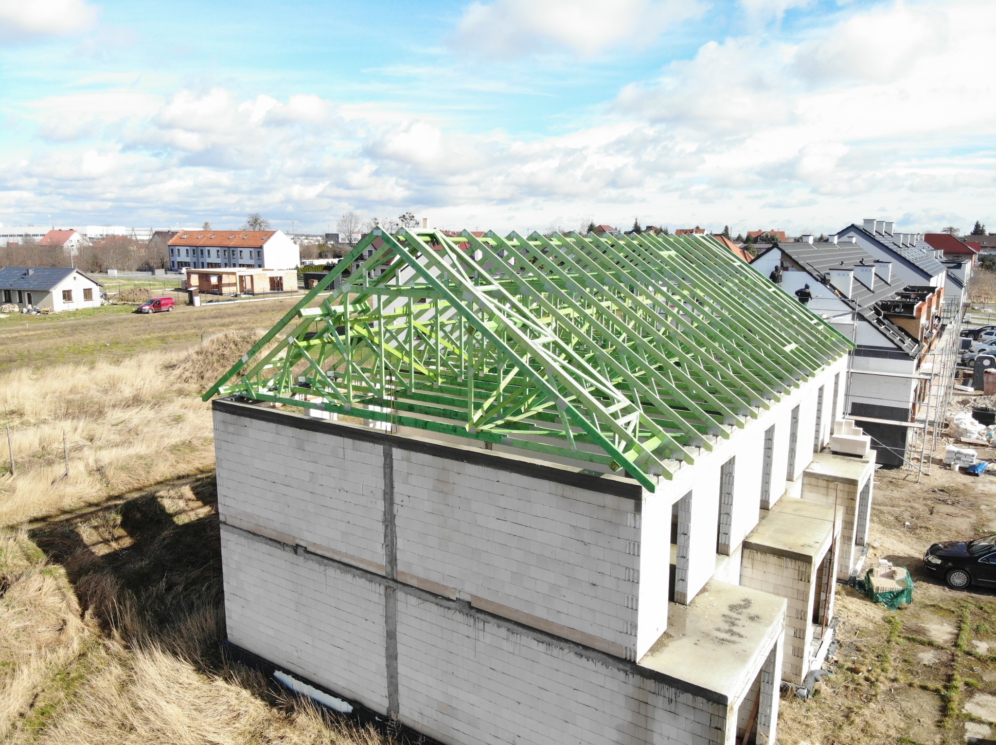 Wznoszenie konstrukcji dachu z impregnowanego drewna na budynku w stanie surowym otwartym z białych bloczków, widok z góry na osiedle domów jednorodzinnych w tle.