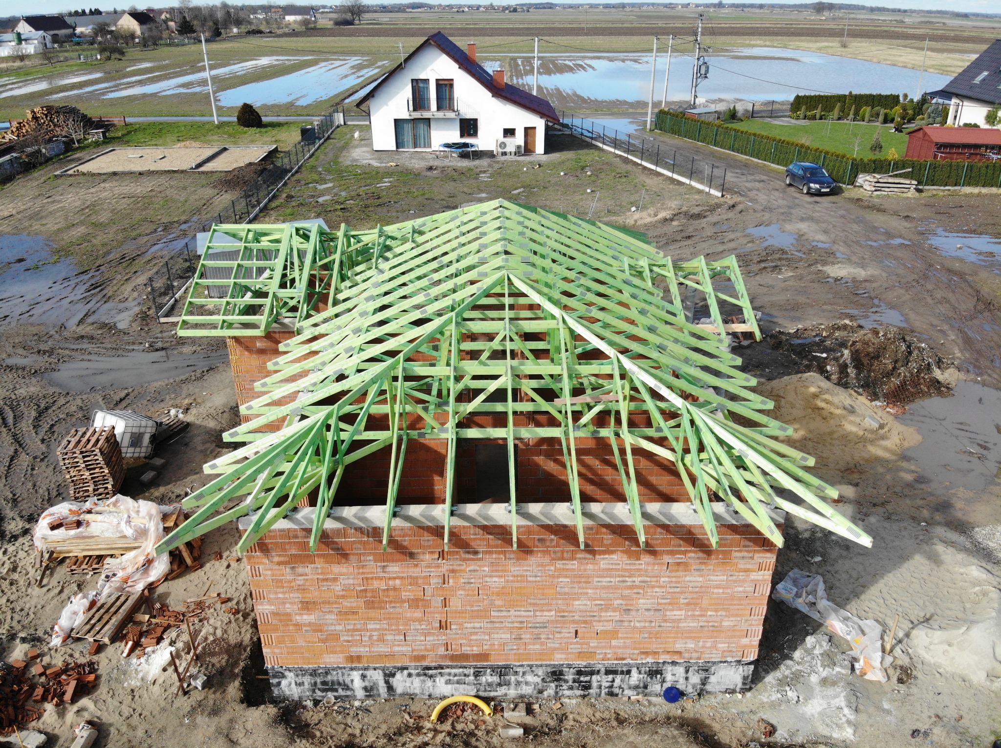 Budowa domu z cegły z zieloną konstrukcją więźby dachowej w trakcie montażu, widok z góry na plac budowy z rozlewiskami wody w tle.