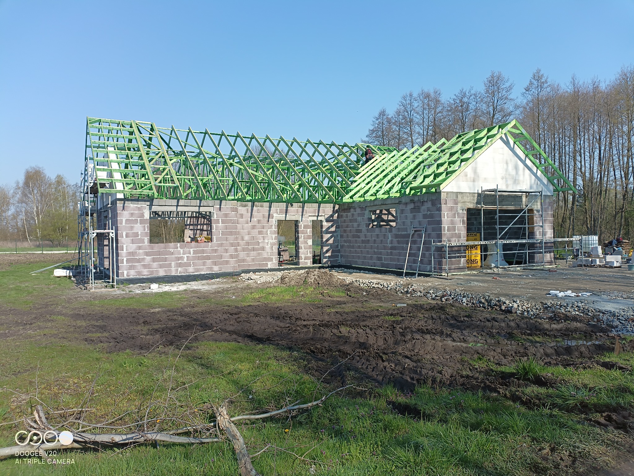 Budowa domu z szkieletową konstrukcją dachu pokrytą zieloną impregnacją, widoczne bloczki betonowe ścian i rusztowania, teren budowy z błotem i trawą.
