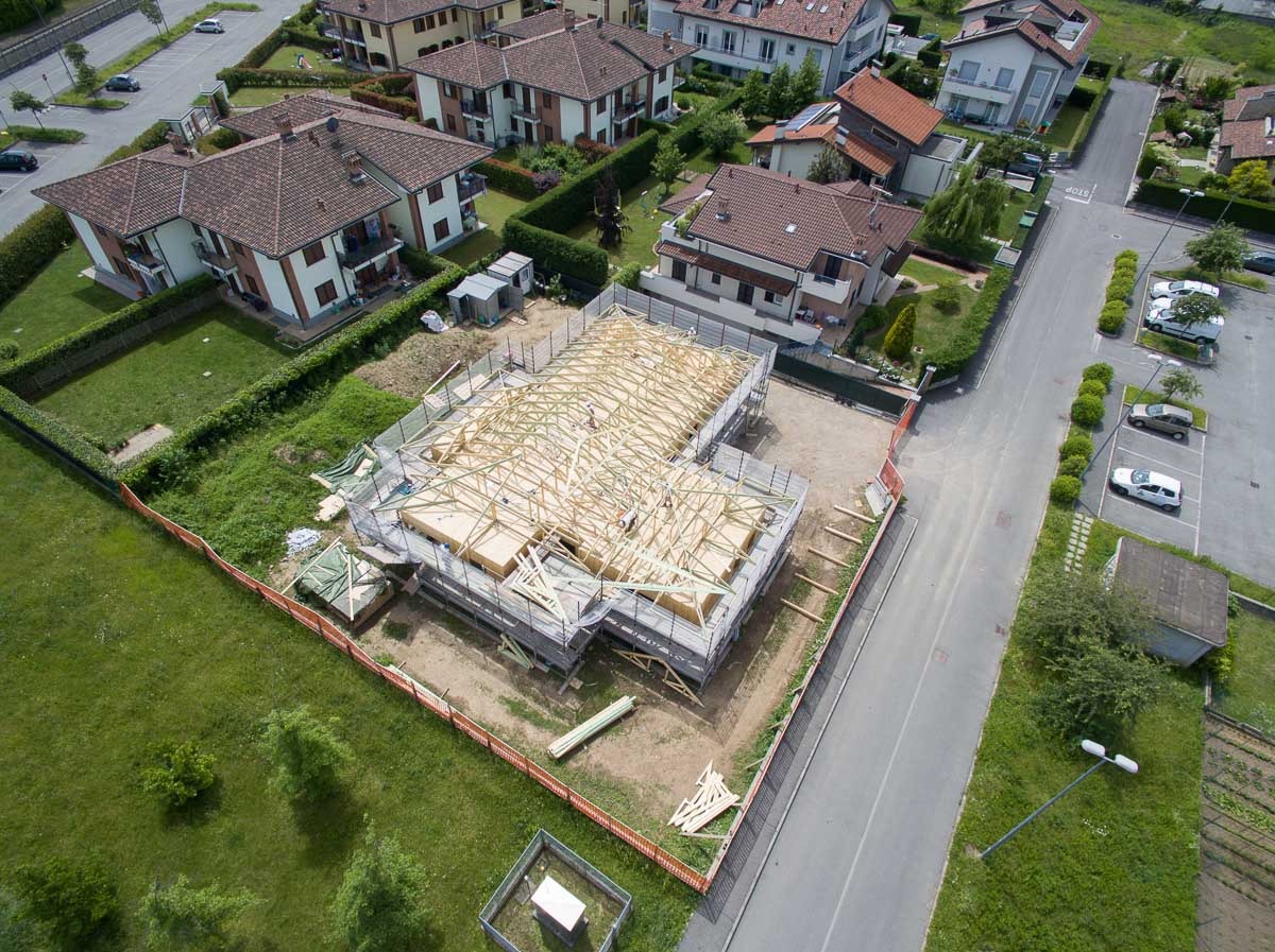 Widok z lotu ptaka na budowę domu z drewnianą konstrukcją dachu, otoczonego ogrodzeniem i zielenią, w tle zabudowa jednorodzinna.